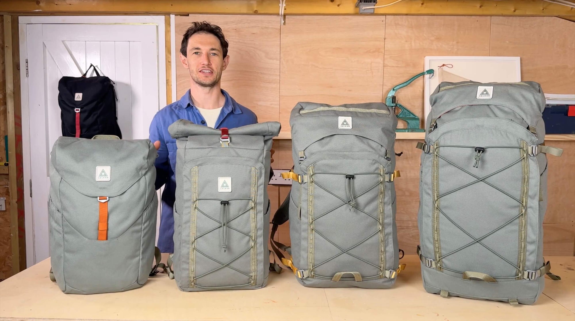 A man stands behind four large gray backpacks of varying sizes arranged on a table in an indoor workshop or studio setting.